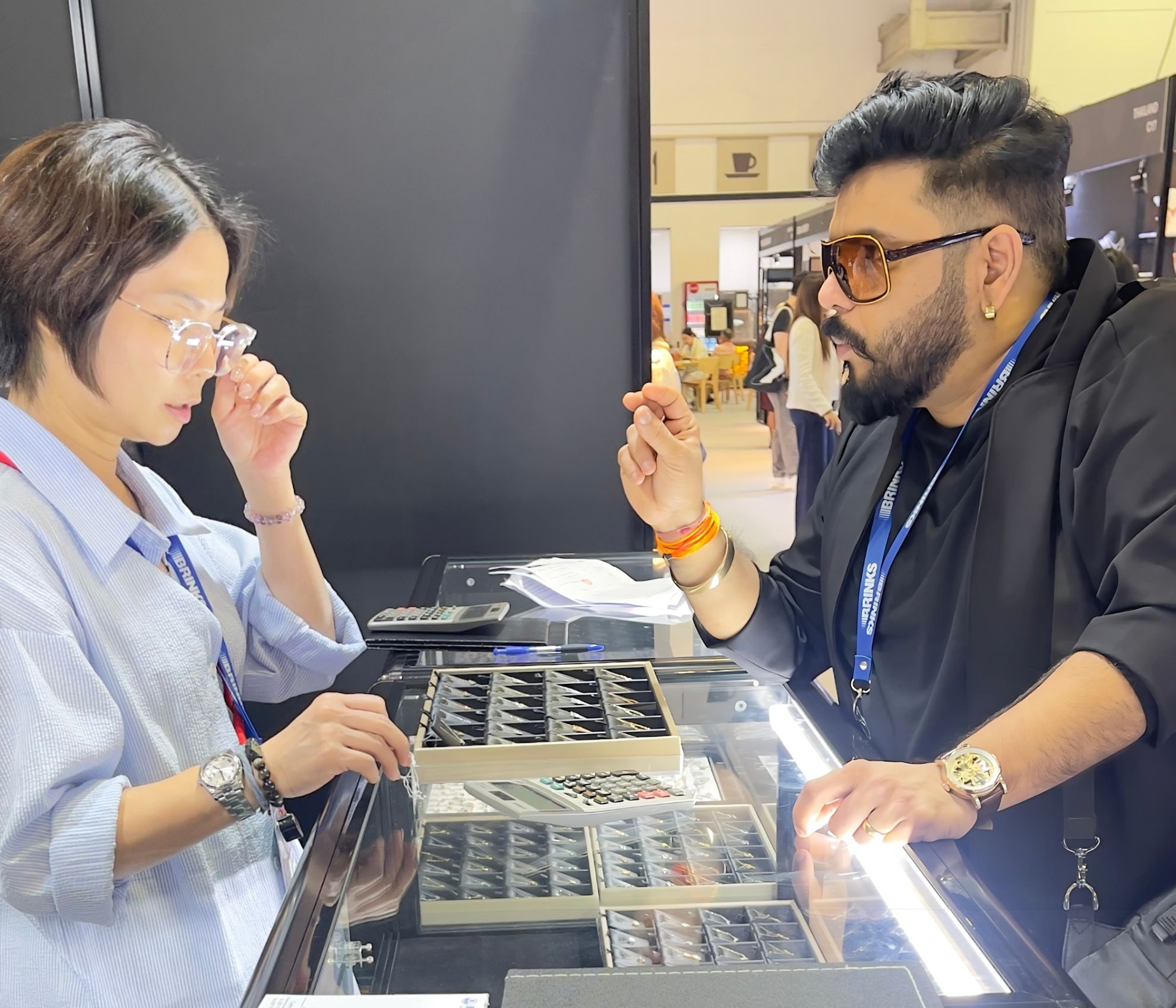 Niraj Choksi and a buyer on opposite sides of a lit display counter at an exhibition stand, examining trays of gold jewellery.