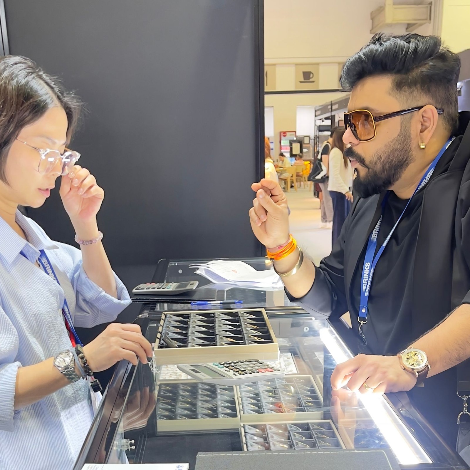 Niraj Choksi and a buyer on opposite sides of a lit display counter at an exhibition stand, examining trays of gold jewellery.